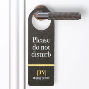 Door handle sign that is hanging on a silver door handle, sign is made out of leather and is printed with "Please do not disturb"