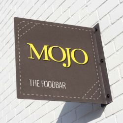 Printed aluminium projecting sign with personalised logo in yellow and white, fixed to a white brick wall with black screw caps