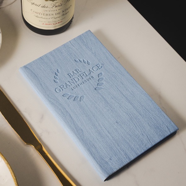 Pale blue Orchard wood-effect bill holder with blind-debossed branding, styled on a restaurant table with wine and gold cutlery